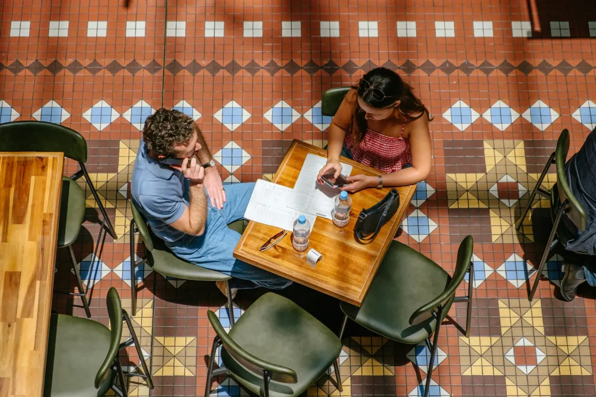 Duas pessoas numa mesa de café sobre um chão de azulejos com padrão, vistas de cima, com ementas e telemóveis