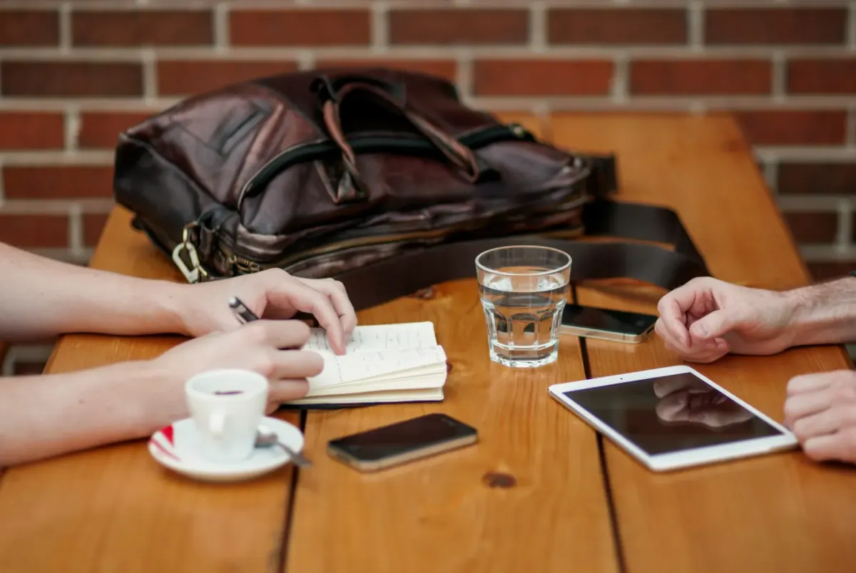 Duas pessoas reunidas numa mesa de café com um caderno, café e um tablet